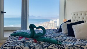 Une chambre d’hôtel avec un lit agrémenté d’une couvre-lit à motifs, une vue sur l’océan et le paysage urbain grâce à une grande fenêtre, et une tête de lit capitonnée.