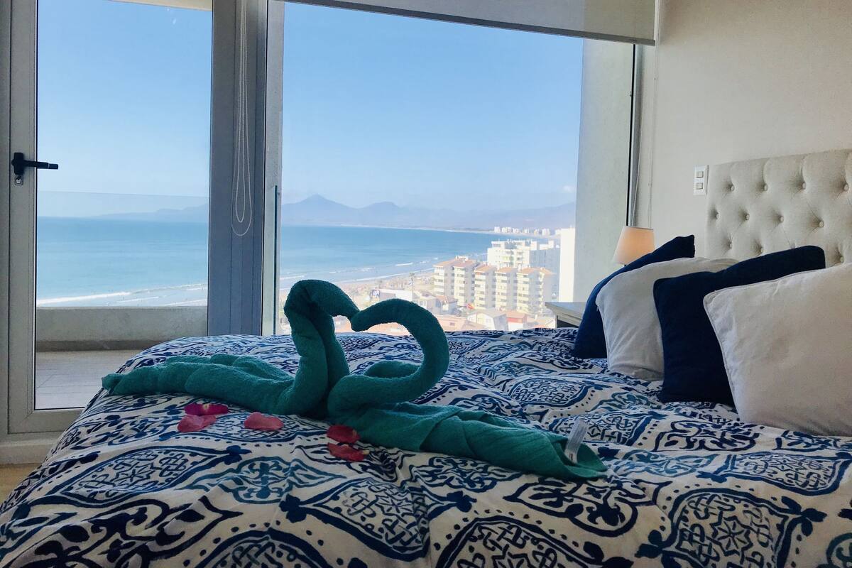 A hotel room with a bed featuring a patterned bedspread, a view of the ocean and cityscape through a large window, and a tufted headboard.