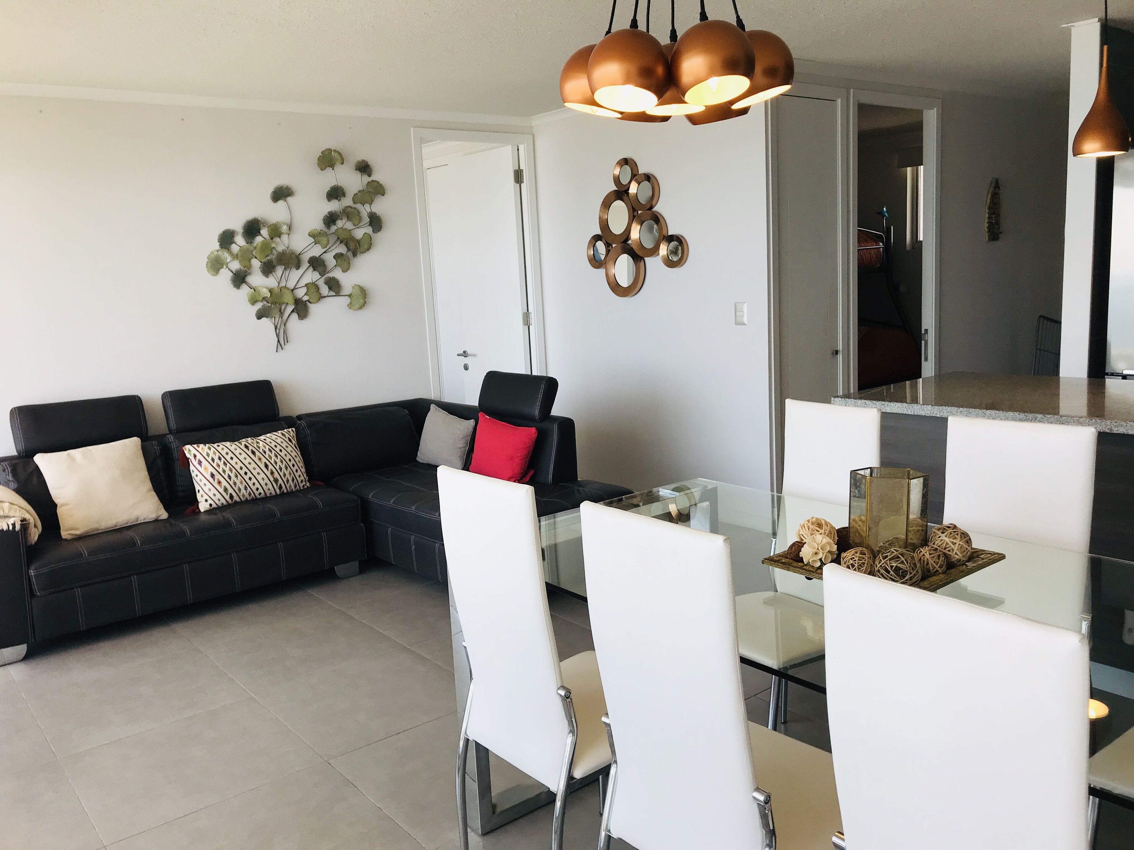 Una sala de estar moderna con un sofá seccional negro, sillas blancas, una mesa de comedor de vidrio y una cocina visible a través de una puerta abierta.