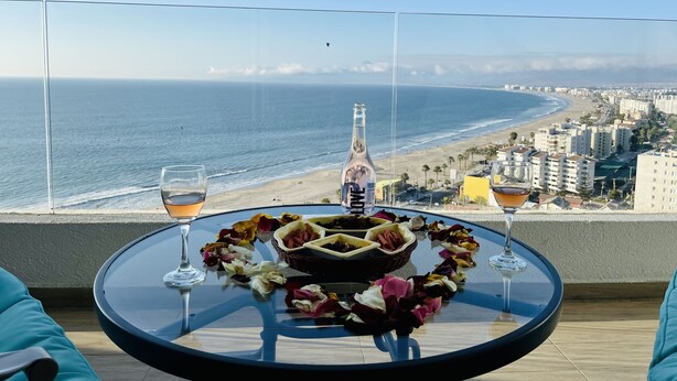 Une table ronde en verre, ornée d’une bouteille, de verres et de pétales de fleurs, est placée sur un balcon donnant sur une plage et sur la ville.