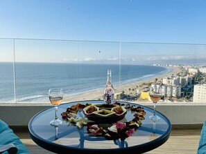 Een ronde glazen tafel met een fles, glazen en bloemblaadjes op een balkon met uitzicht op een strand en stadsbeeld.