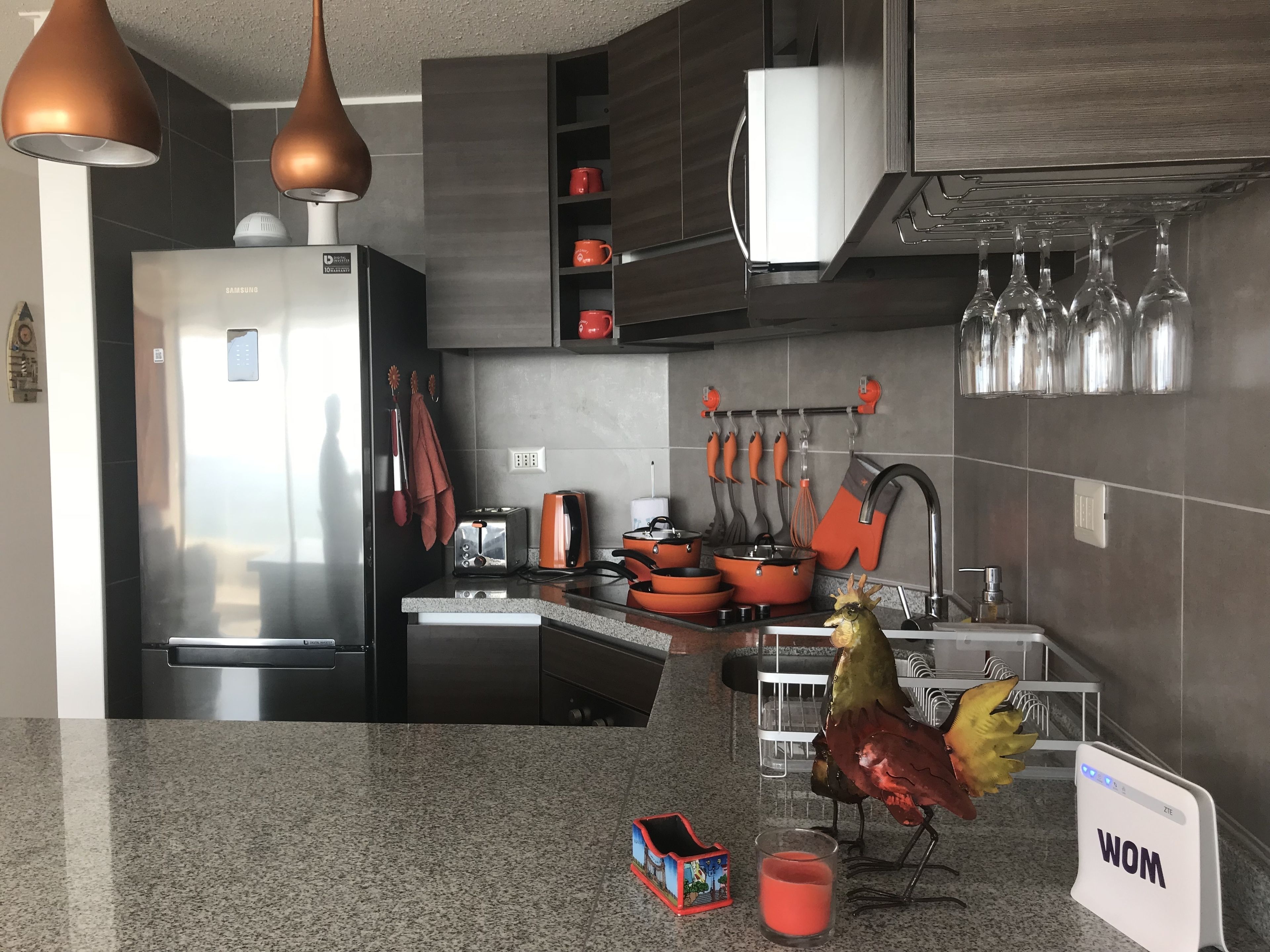 Una cocina moderna con un refrigerador de acero inoxidable, un tostador y una cafetera. Hay ollas y sartenes naranjas en la encimera, y una decoración de gallo en la pared.