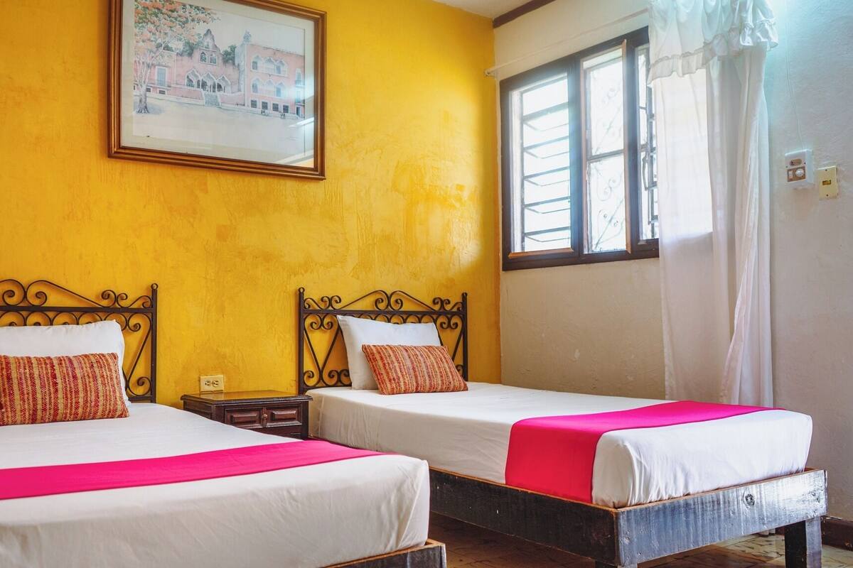 Two beds with metal frames, white bedding, and red accents, in a room with yellow walls and a window with white curtains.