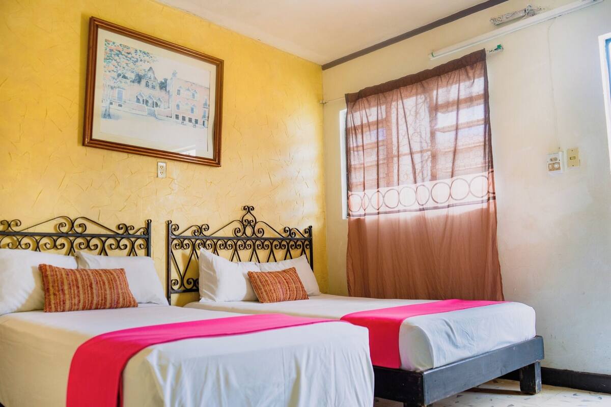 A room with two beds, a framed picture on the wall, a window with curtains, and a thermostat on the wall.