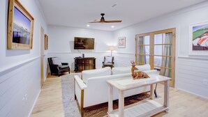 A living room with a white sofa, a wooden coffee table, and a ceiling fan. There are framed pictures on the walls and a window with a view of a bridge. - Exclusive Property Just Steps from History & Culture (Charleston)