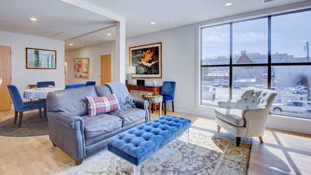 A modern living room with a sofa, ottoman, and armchairs, a dining area with a table and chairs, and large windows offering a city view. - Exclusive Property Just Steps from History & Culture (Charleston)