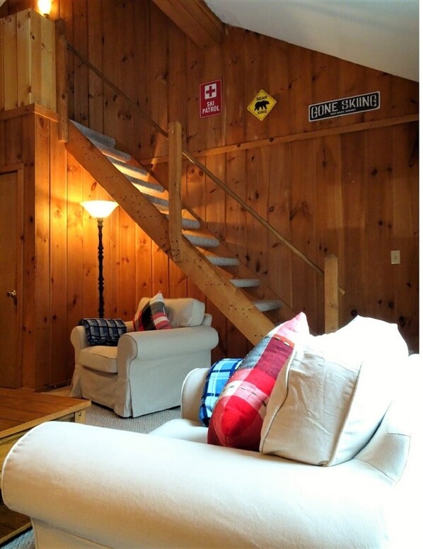 Interior de madera con una escalera, un sofá y una lámpara. Señales indican presencia de patrulla de esquí y advertencia por cruce de oso.