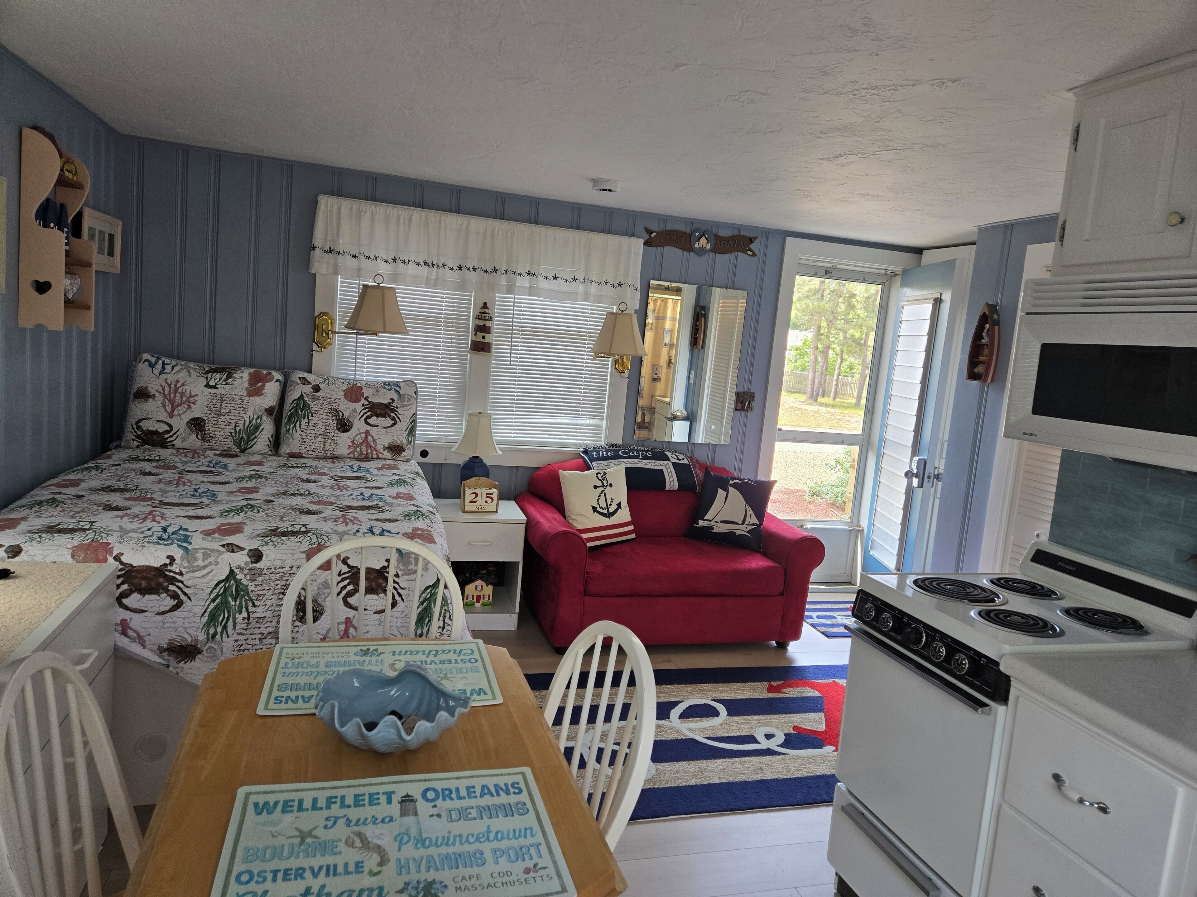 A cozy room with a bed, a red chair, a table with a placemat, a stove, and a window with blinds.