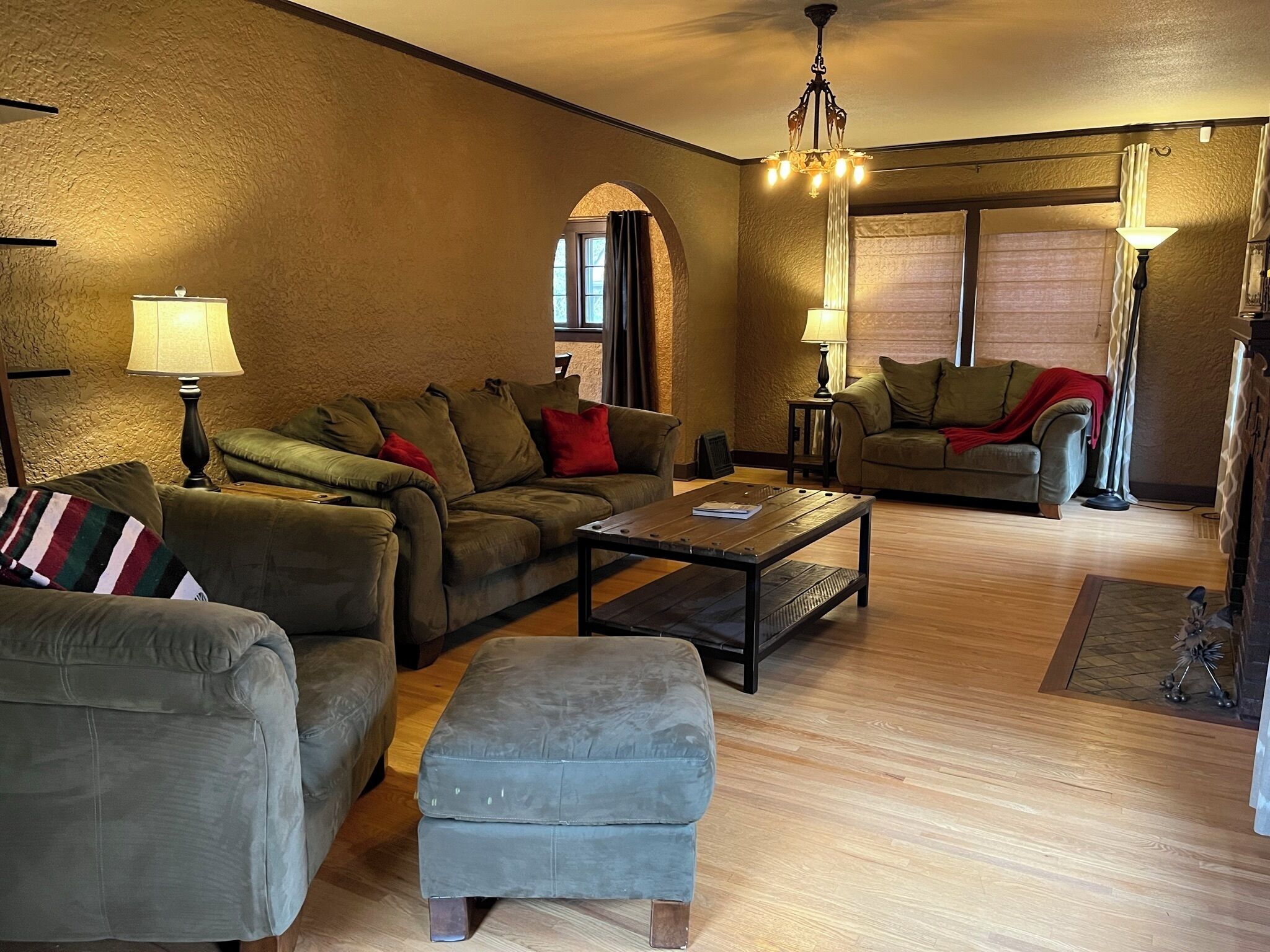 A cozy living room with a sectional sofa, an ottoman, and a coffee table. There are two lamps and a chandelier. The room has wooden flooring and a large window with curtains.