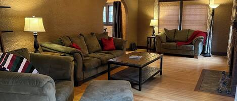 A cozy living room with a sectional sofa, an ottoman, and a coffee table. There are two lamps and a chandelier. The room has wooden flooring and a large window with curtains.