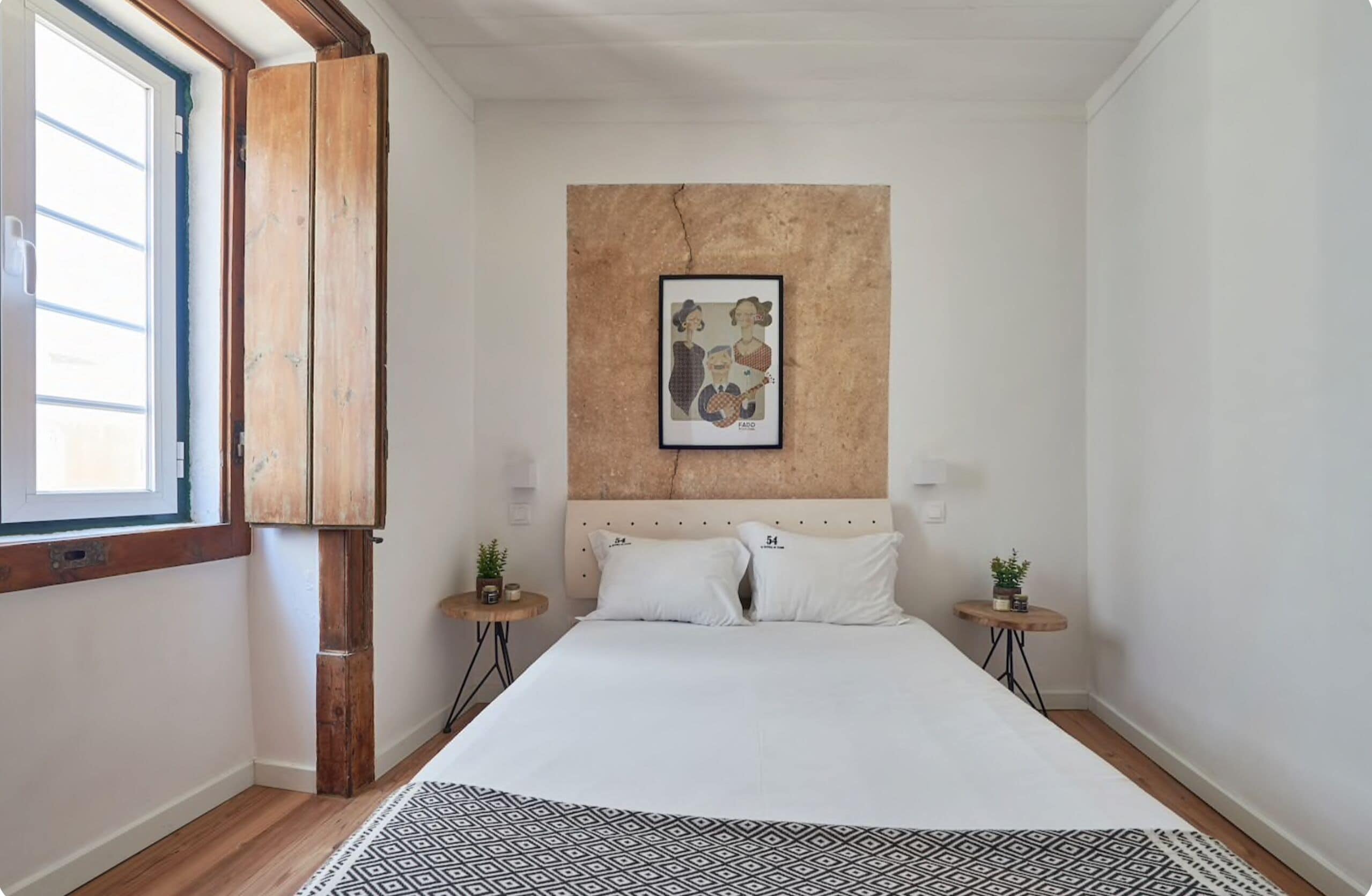 A bedroom with a large bed, a wooden wardrobe, a framed artwork on the wall, two bedside tables with plants, and a window with blinds.