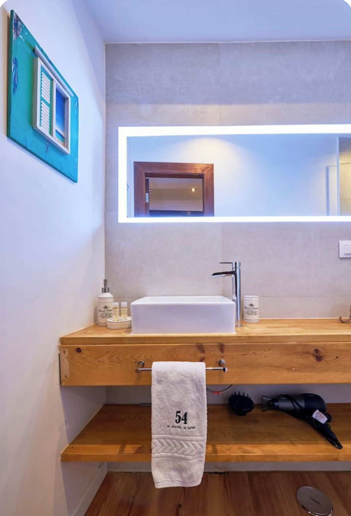 A modern bathroom with a wooden vanity, a white sink, and a large mirror with integrated lighting.