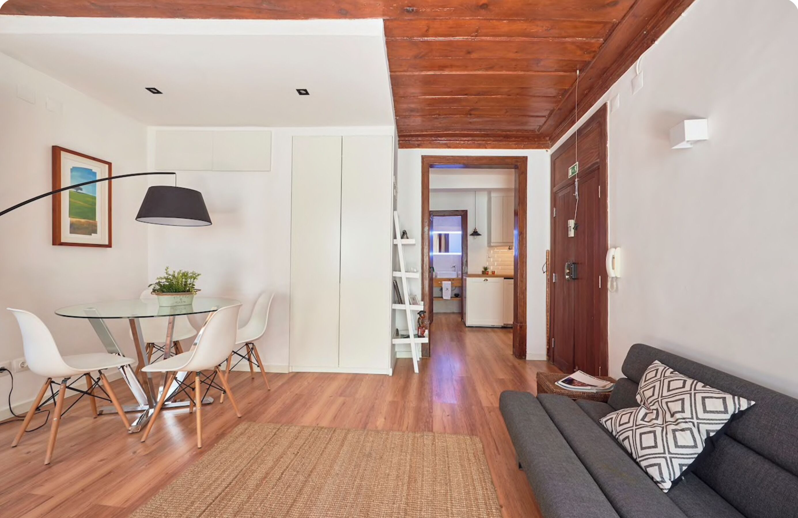 Ein modernes Wohnzimmer mit einem Glastisch, weißen Stühlen, einem grauen Sofa und einer Holzbalkendecke.