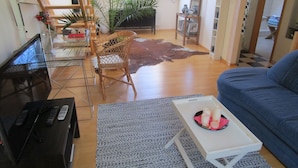 A living room with a sofa, a coffee table with candles, a TV, and a laptop. There is a chair, a rug, and a plant.