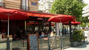 Un café en plein air avec des parasols rouges, des tables en verre et un menu écrit à la craie sur un tableau noir.