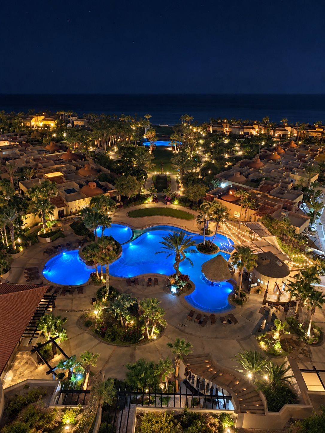 A resort with multiple swimming pools, palm trees, and a view of the ocean at night.