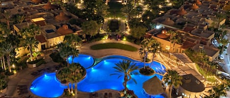 A resort with multiple swimming pools, palm trees, and a view of the ocean at night.