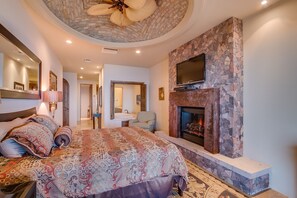 A bedroom with a large bed, a stone fireplace, a television, and a ceiling fan.