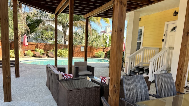A covered patio area with wicker furniture and a glass table, overlooking a swimming pool and a yellow building. - F1, Downtown St. Augustine, POOL, beaches, free onsite parking! (Saint Augustine)