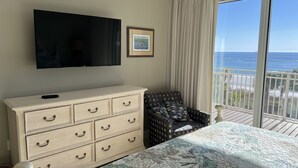 A bedroom with a bed, a dresser, a chair, a TV, and a balcony with a view of the ocean.
