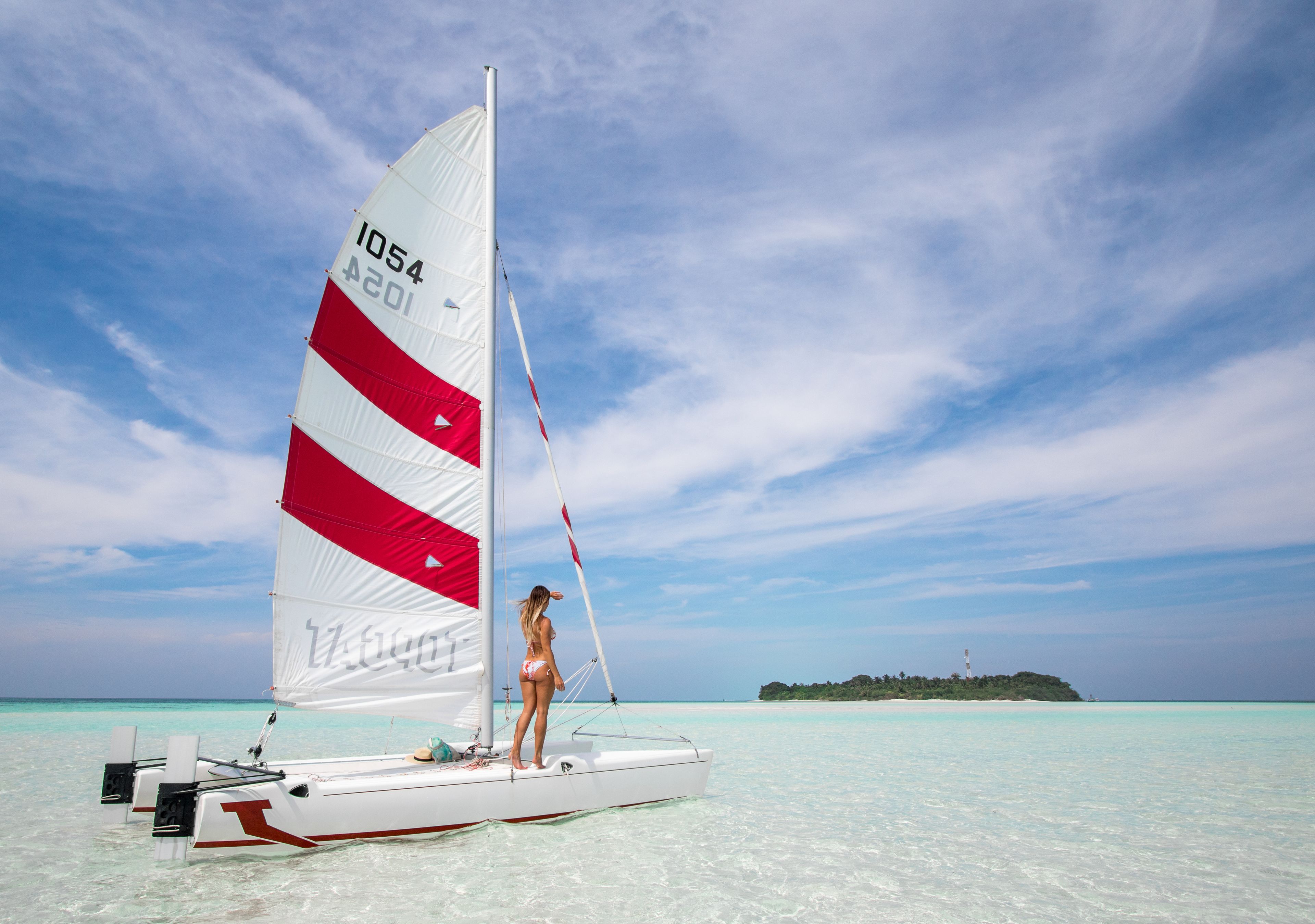 A sailboat with a woman standing on the deck, a clear blue sky, and a distant island.