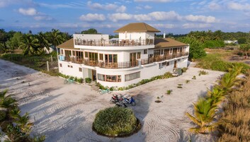 A two-story building with a balcony, surrounded by sand and palm trees.