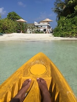 A kayak with a yellow paddle, a sandy beach, and a building with a balcony in the background.