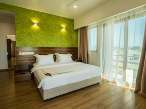 Habitación de hotel con una cama grande, cabecera de madera, pared verde como detalle decorativo, dos lámparas de noche y vistas a la vegetación a través de cortinas translúcidas.