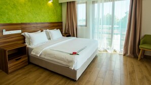 A hotel room with a large bed, wooden headboard, a nightstand, a green chair, and a view of the outdoors through sheer curtains.