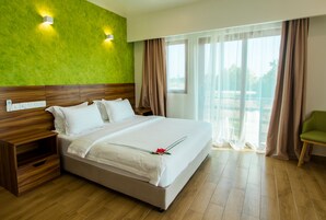 A hotel room with a large bed, wooden headboard, a nightstand, a green chair, and a view of the outdoors through sheer curtains.