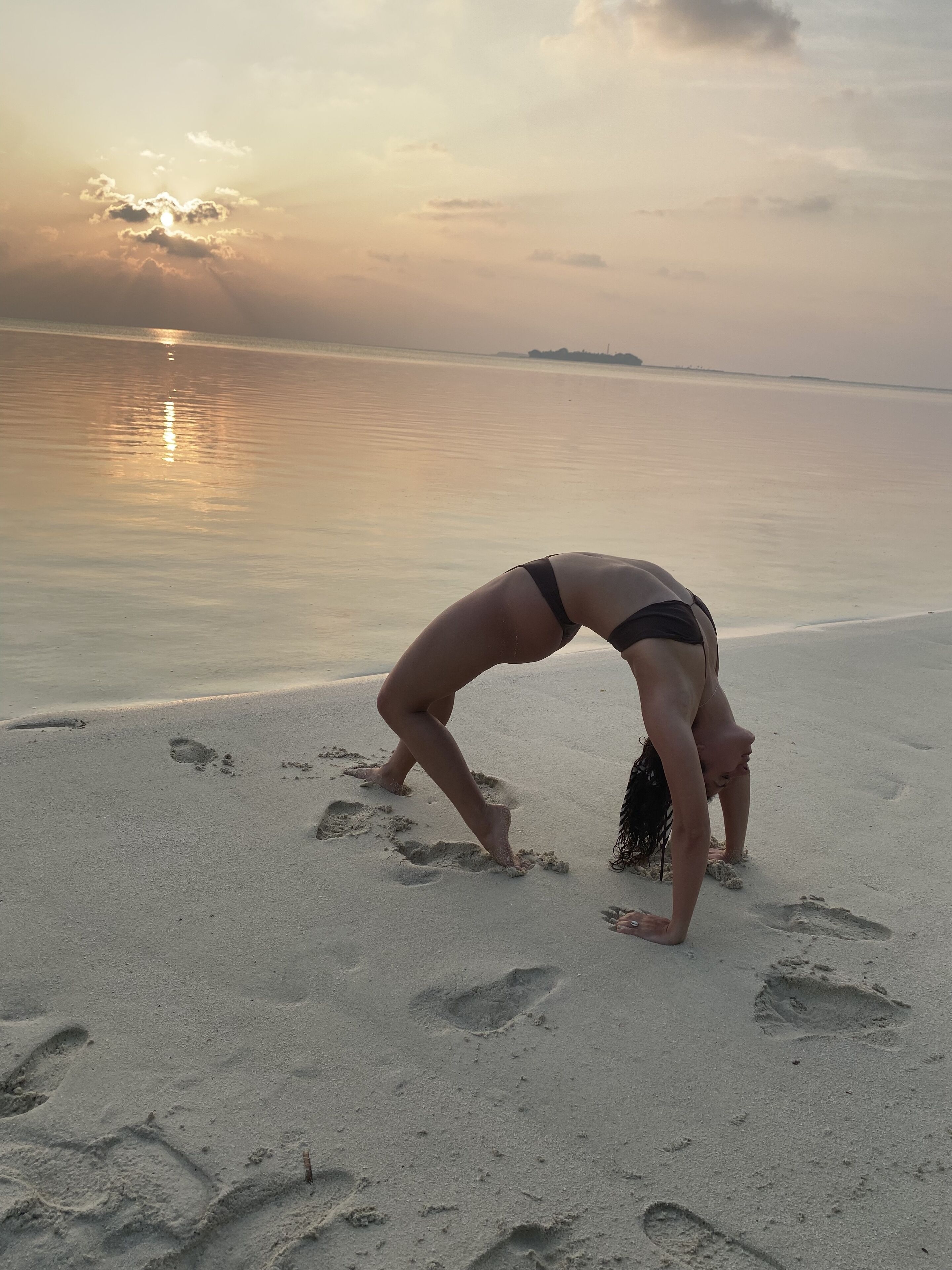 A person performing a yoga pose on a sandy beach with footprints, a calm sea, and a sunset in the background.