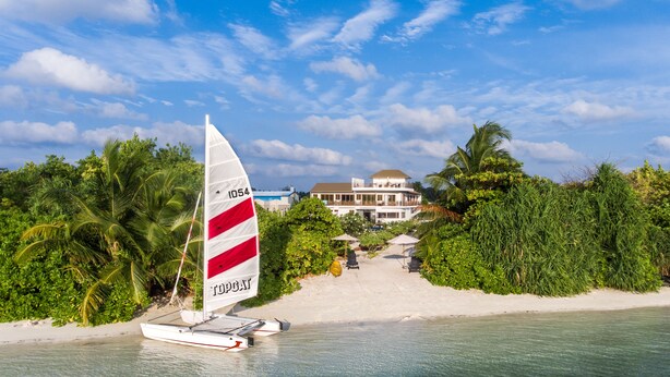 A sailboat on a sandy beach with lush greenery and a building in the background.