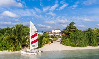 A sailboat on a sandy beach with lush greenery and a building in the background.