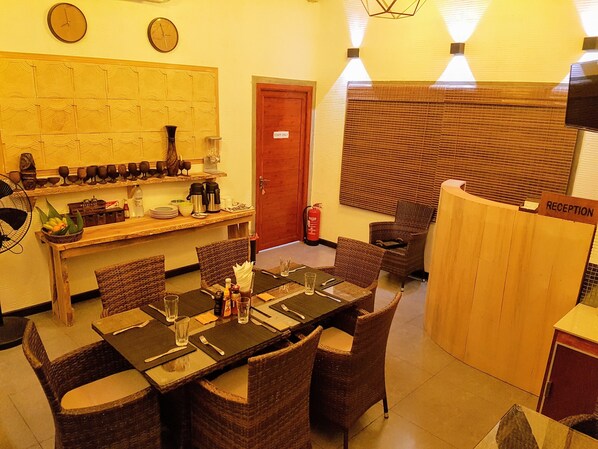 A dining area with a long table set for four, surrounded by wicker chairs. There is a reception desk to the right and a wall with clocks above it.