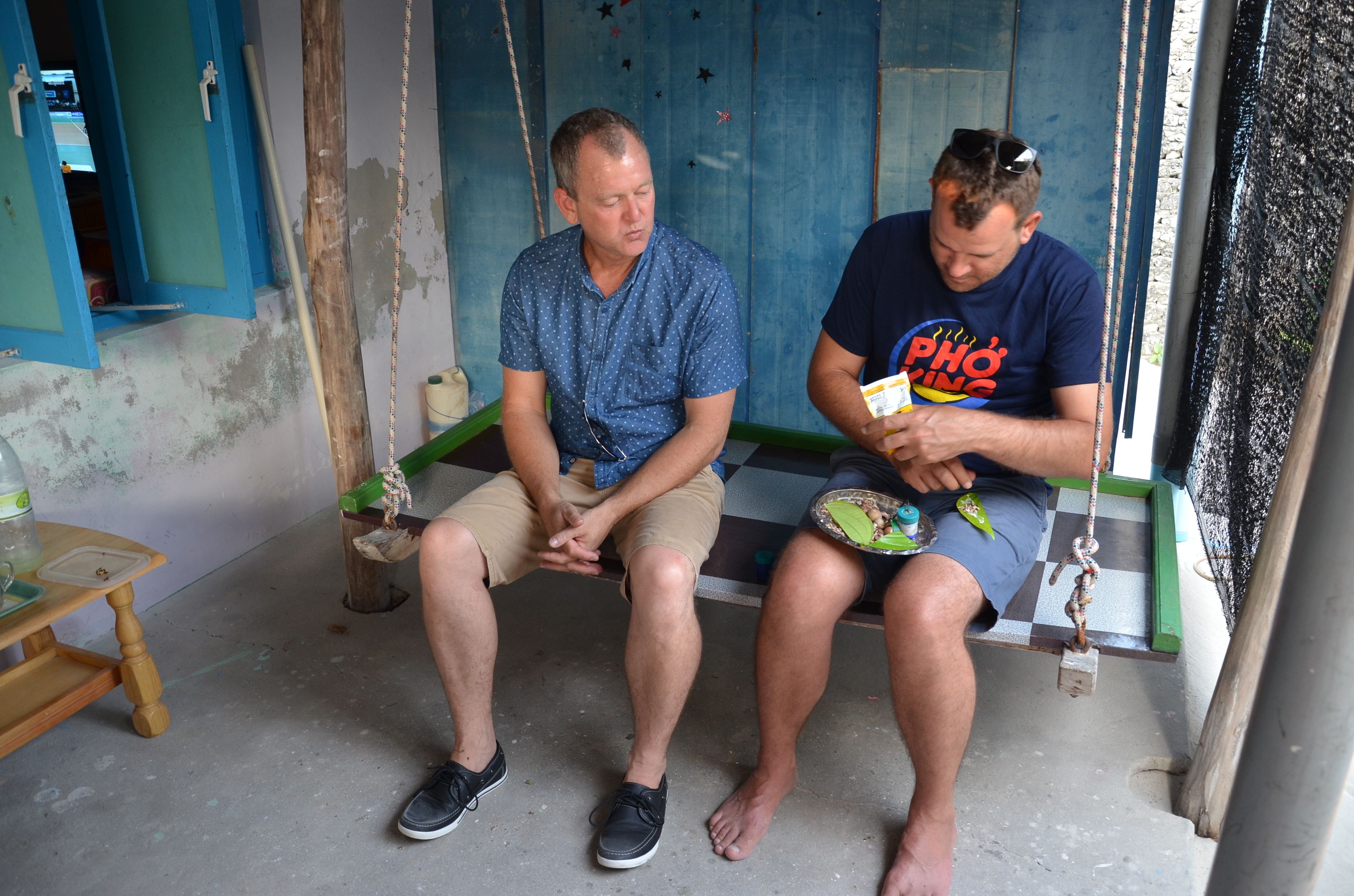 Two men sitting on a swing in a room with a blue wall and a checkered floor.