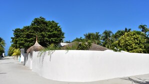 Um edifício de paredes brancas com fachada curva, uma estrutura com telhado de palha e vegetação exuberante.