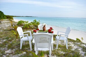 A beachside table with white chairs and a vase of flowers, overlooking a sandy beach and calm sea.
