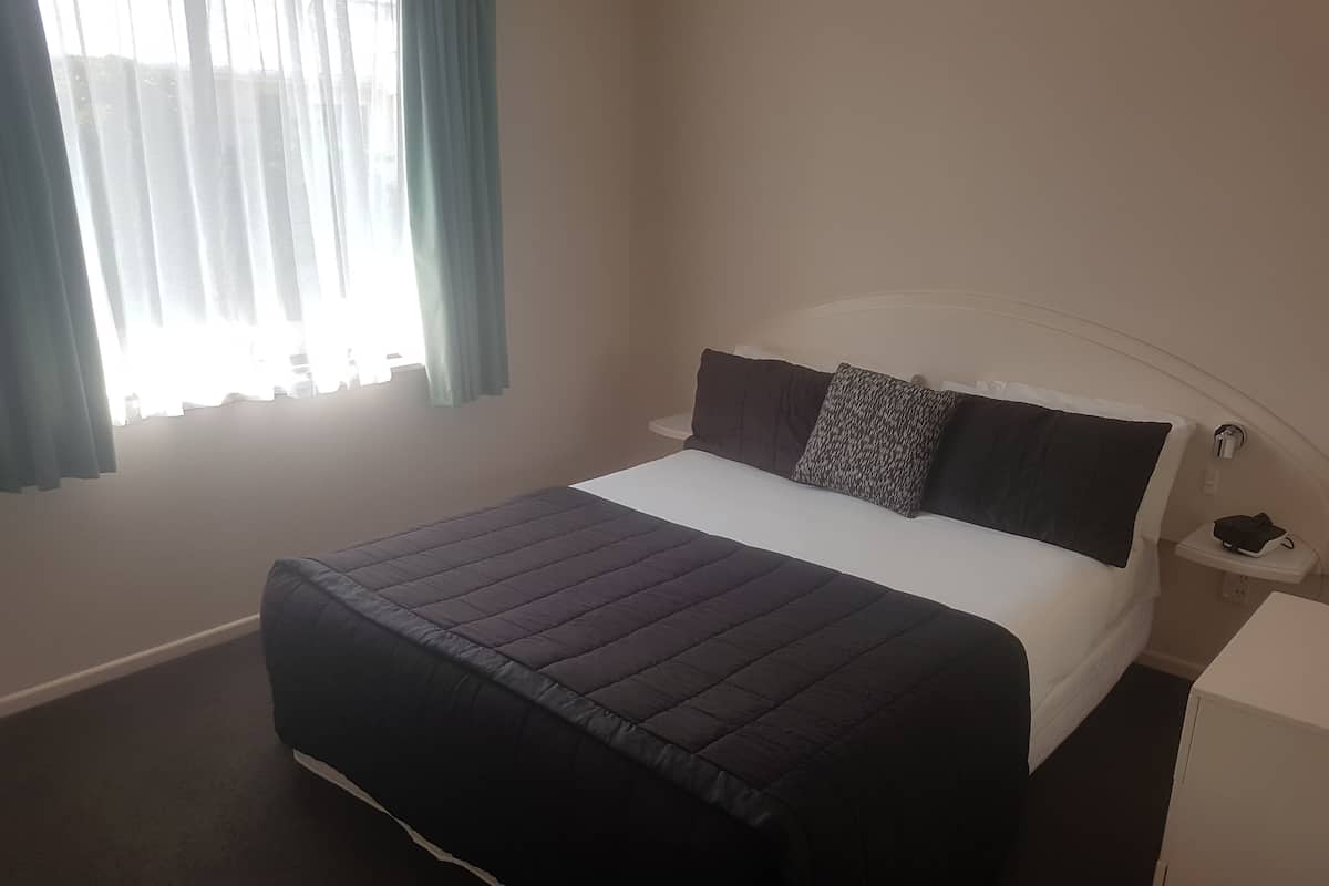 A neatly made bed with a dark-colored duvet and pillows, a bedside table with a phone, and a window with curtains.