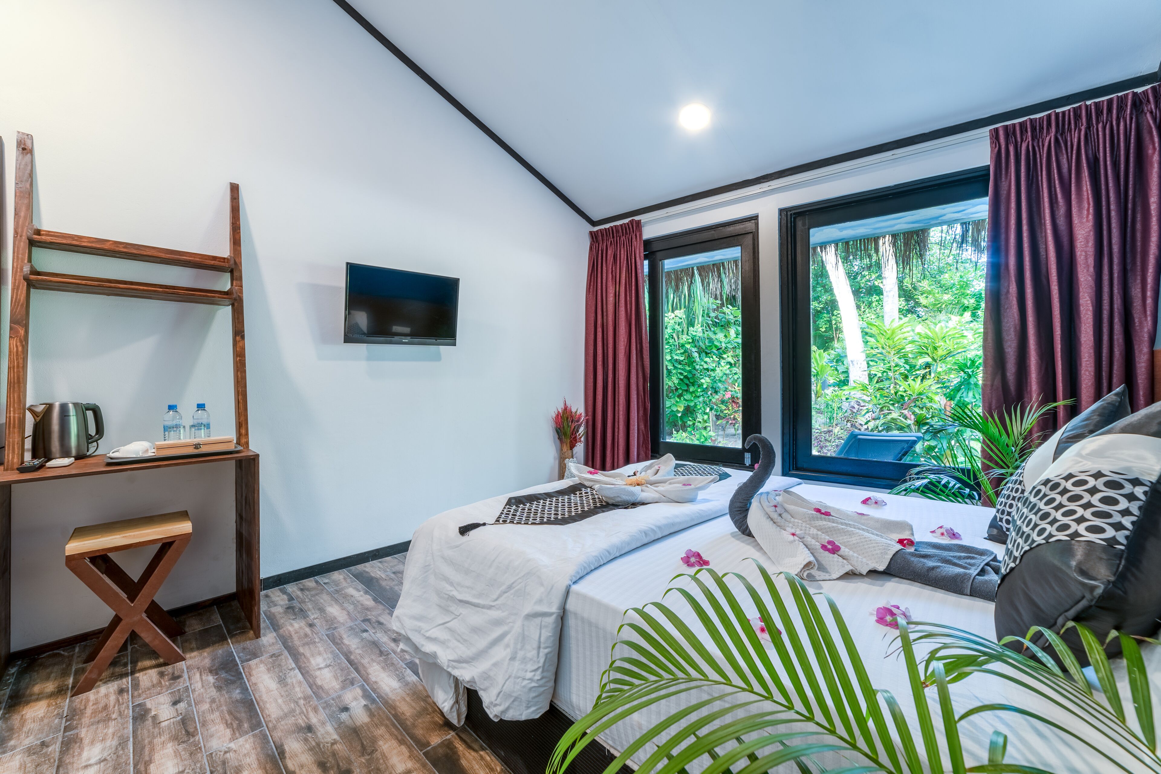 Habitación moderna con cama, escritorio y televisor. Hay plantas y una ventana con cortinas.