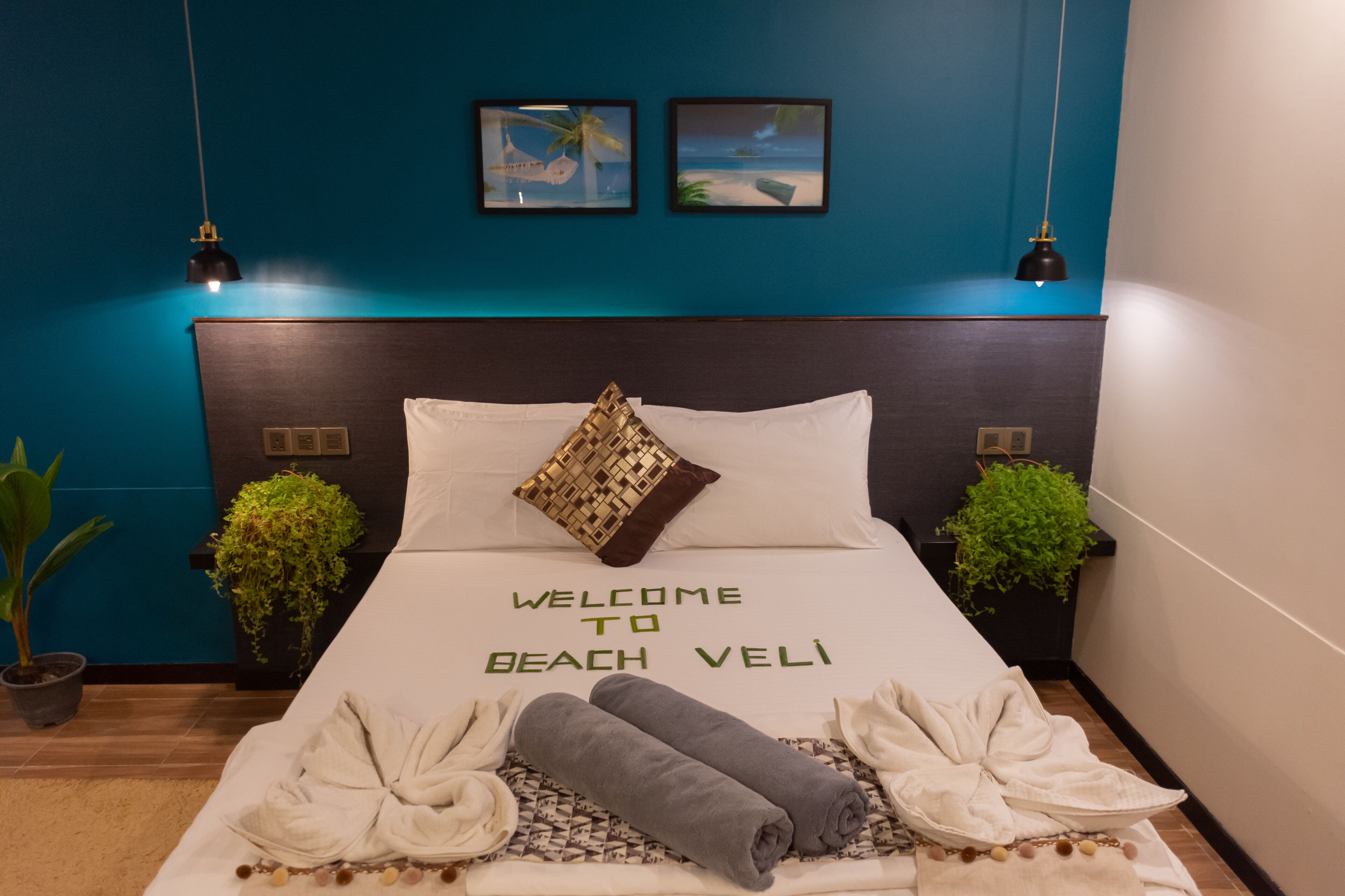Una cama impecable con un cartel que dice "Welcome to Beach Veli", dos pequeñas macetas con plantas y vista al océano a través de la ventana.