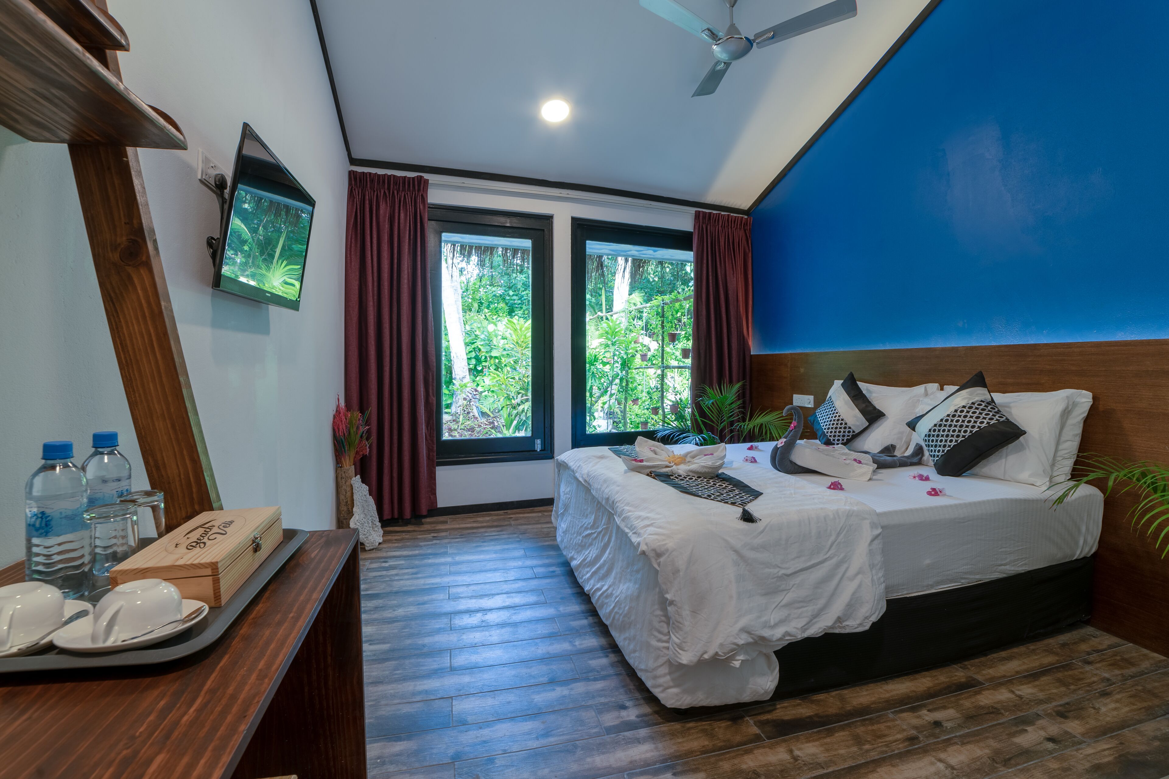Quarto com cama, escrivaninha com objetos sobre uma bandeja, televisão na parede e janela com cortinas.