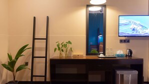 Habitación de hotel con un escritorio de madera, un cubo de basura pequeño, una planta en maceta, una escalera y un televisor de pantalla plana montado en la pared.