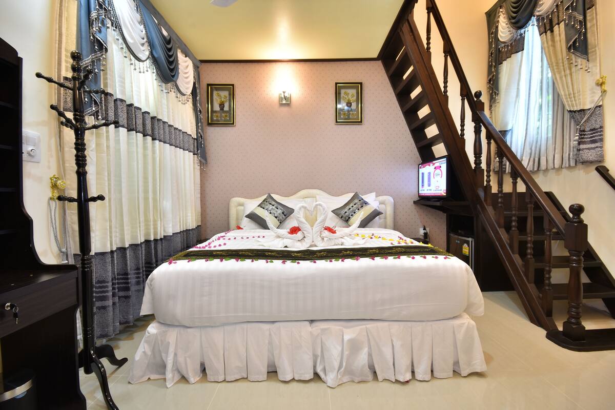 Una cama grande con una colcha blanca, dos almohadas y un cabecero decorativo. Se observa una escalera de madera a la derecha y un televisor fijado a la pared sobre una pequeña mesa junto a la cama.