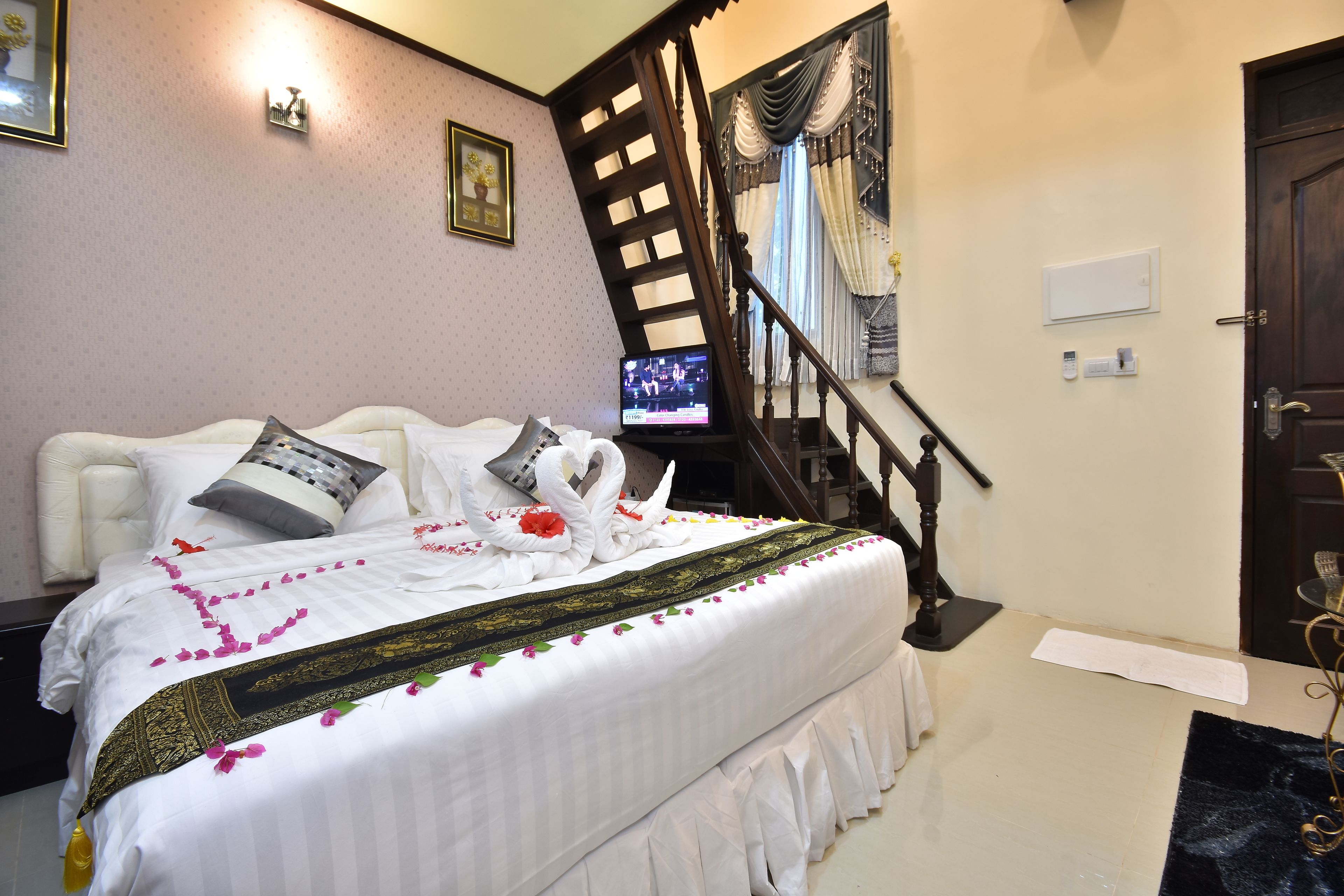 A neatly made bed with a white and gold bedspread, a pair of swan-shaped pillows, and a bouquet of pink flowers.