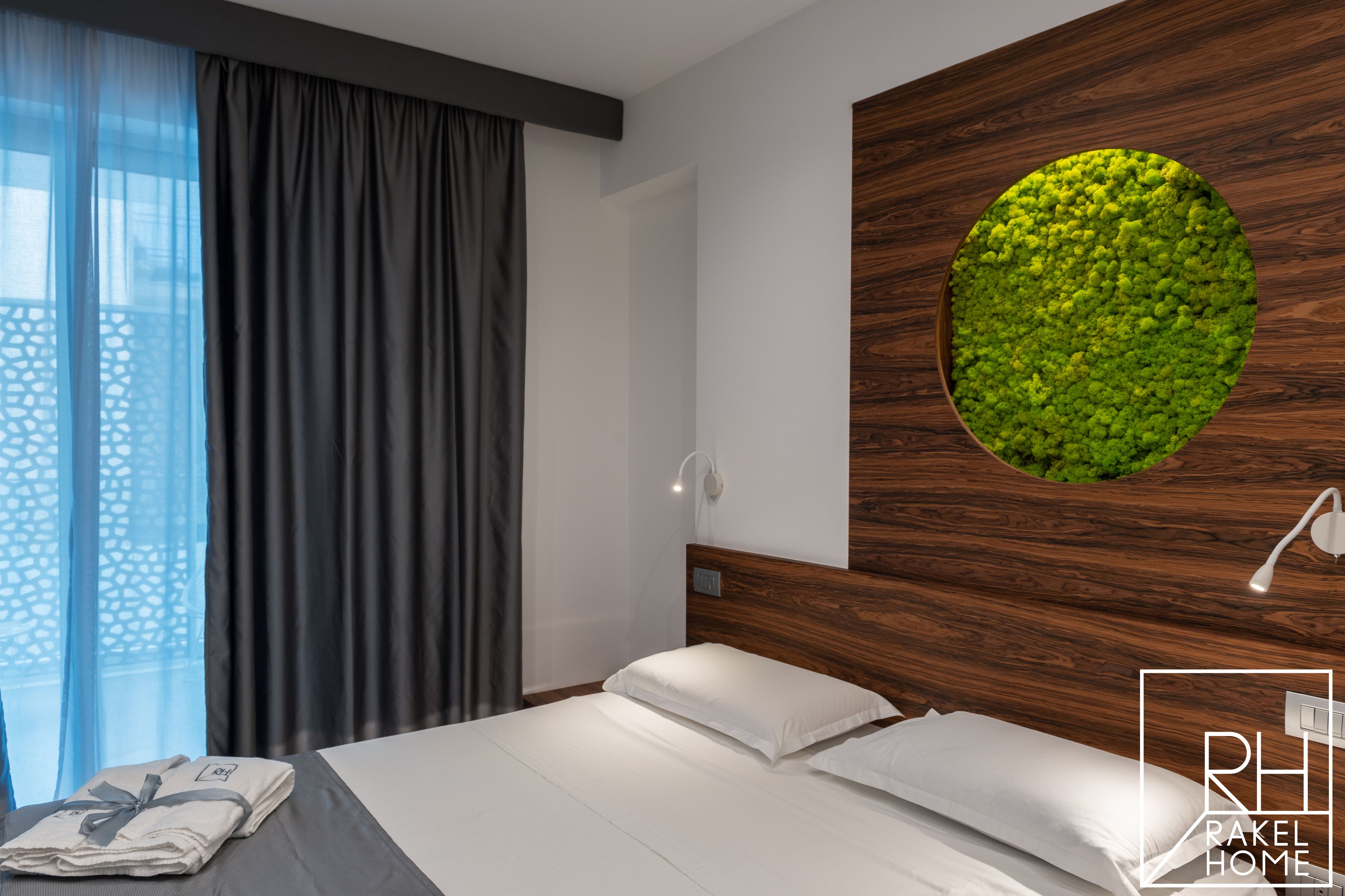A modern hotel room with a neatly made bed, a large window with sheer curtains, and a unique wall feature with a circular green moss-like design.