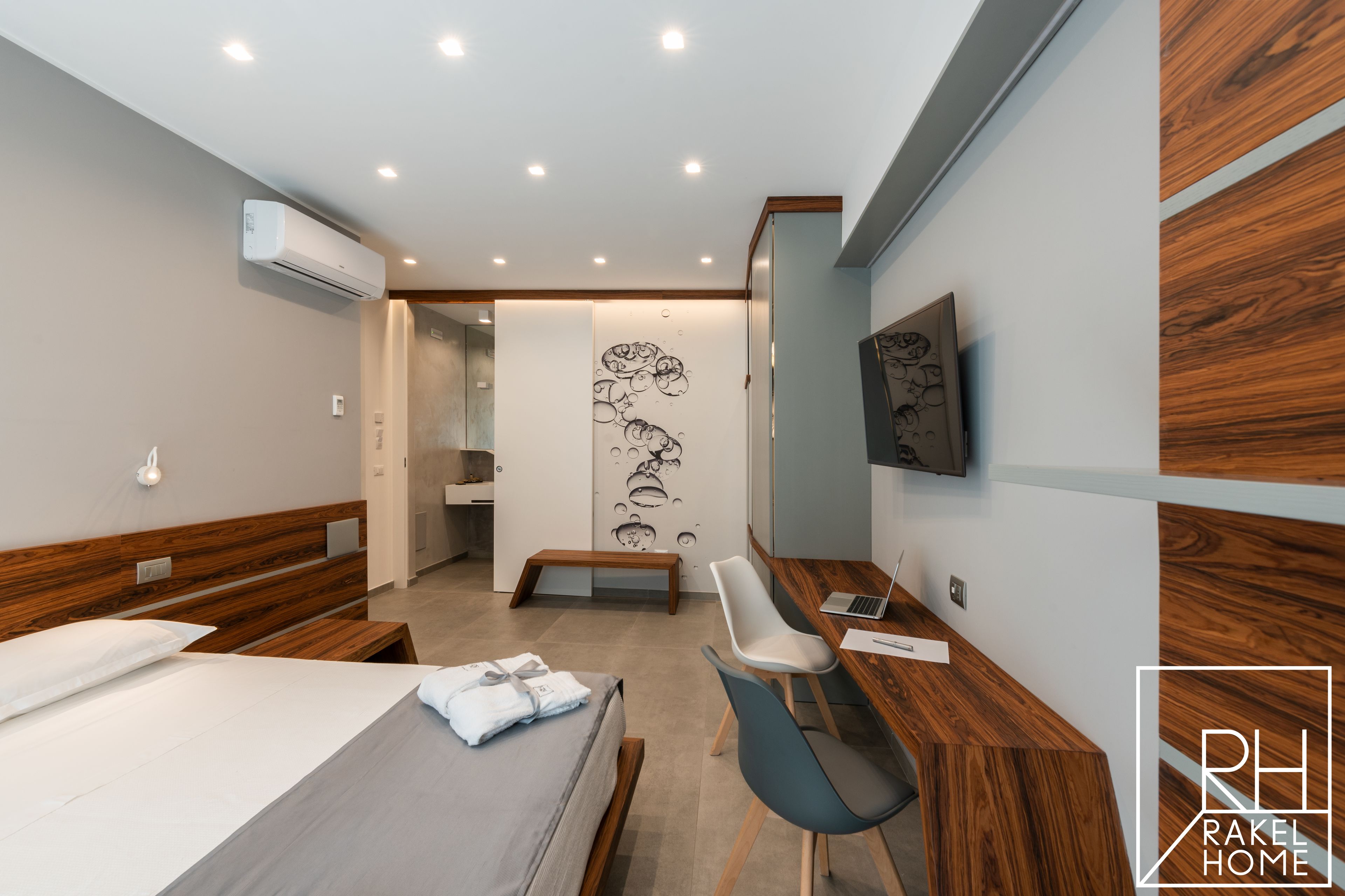 A modern hotel room with a bed, a desk with a laptop, a chair, and a wall-mounted TV.