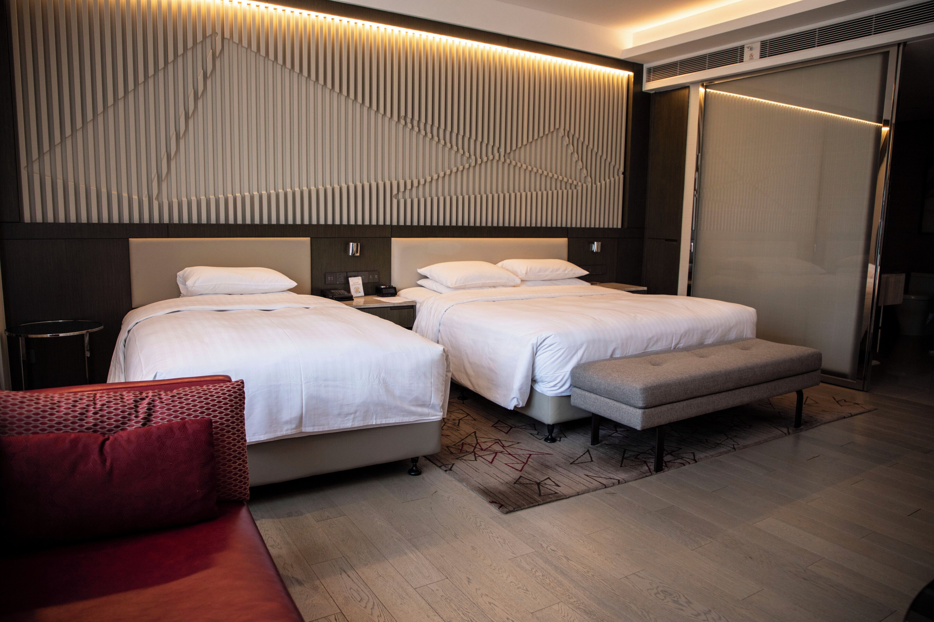 A modern hotel room with two beds, a red sofa, and a wooden floor.