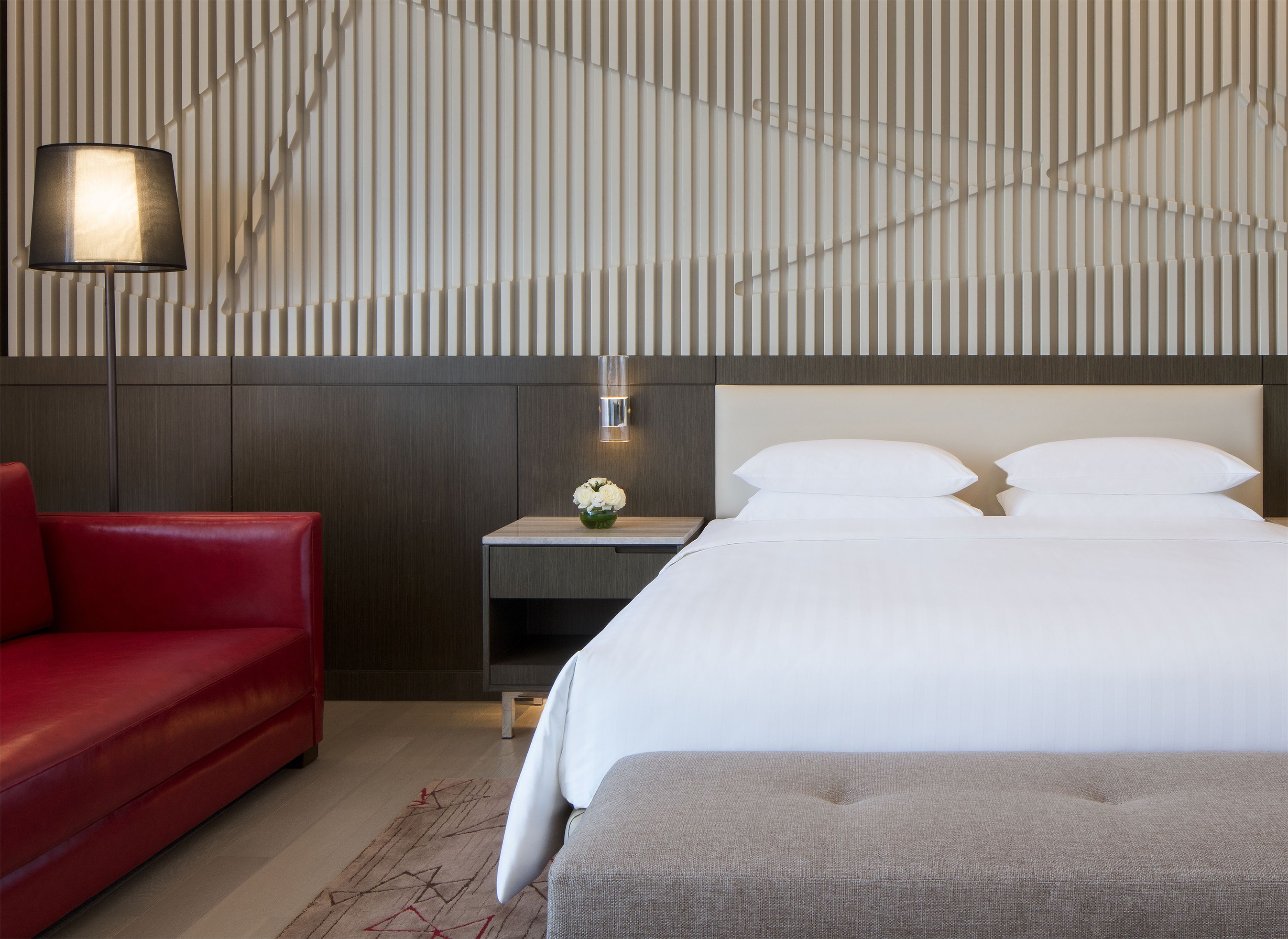 Habitación de hotel con cama, sofá rojo, mesita de noche con un jarrón de flores y una pared con líneas verticales.
