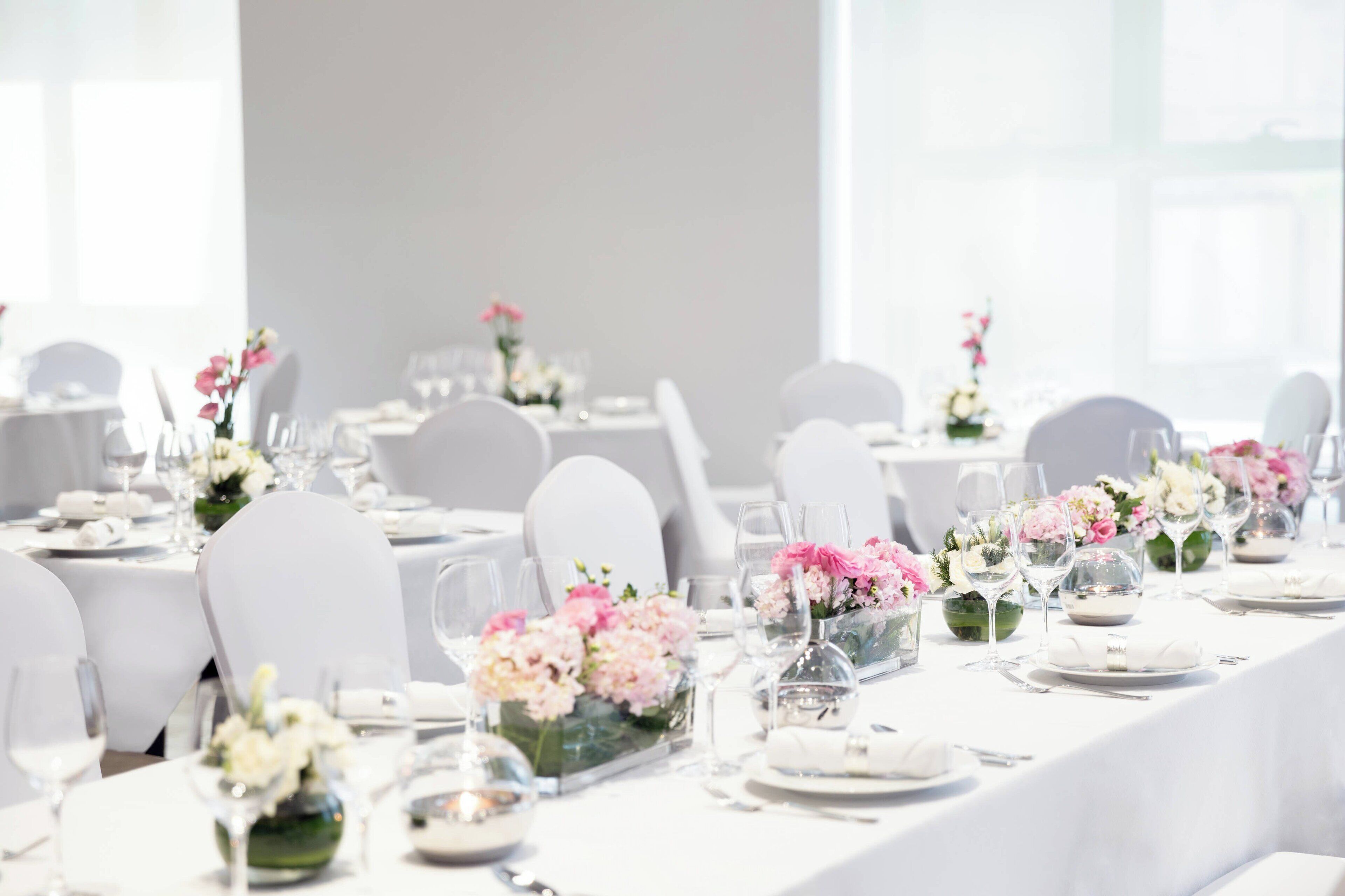 Salão de festas com mesas brancas dispostas para um evento formal, cada uma com um arranjo floral e copos de vidro transparente.