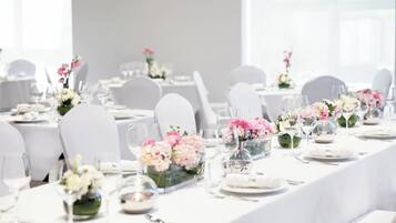 Salão de festas com mesas brancas dispostas para um evento formal, cada uma com um arranjo floral e copos de vidro transparente.